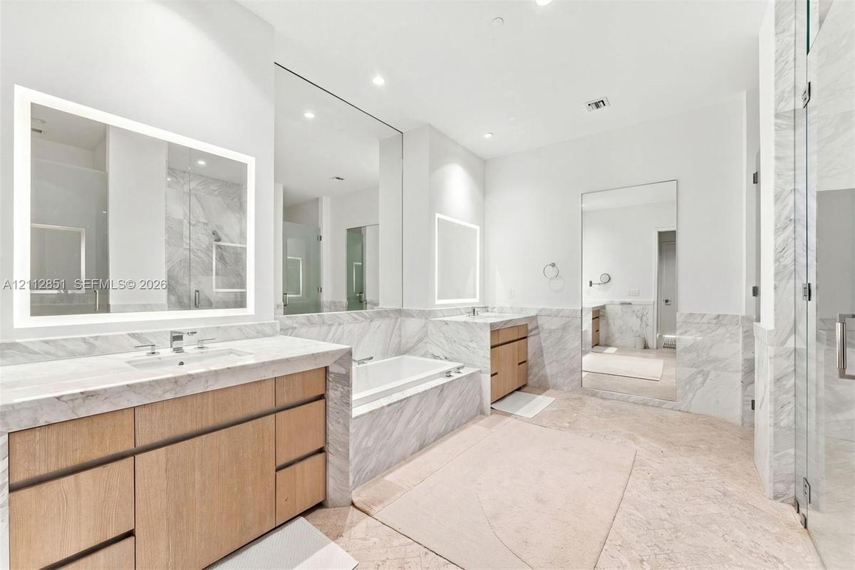 Dual wood-finish vanities with stone countertops, built-in soaking tub, stone tile flooring, and illuminated wall-mounted mirrors