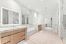 Dual wood-finish vanities with stone countertops, built-in soaking tub, stone tile flooring, and illuminated wall-mounted mirrors