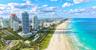 Aerial perspective of a coastal cityscape featuring high-rise buildings, extensive sand coastline, and turquoise ocean waters