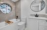 Bathroom featuring large-format tile surround, circular accent window, white vanity with integrated basin, matte black fixtures, and oval wall-mounted