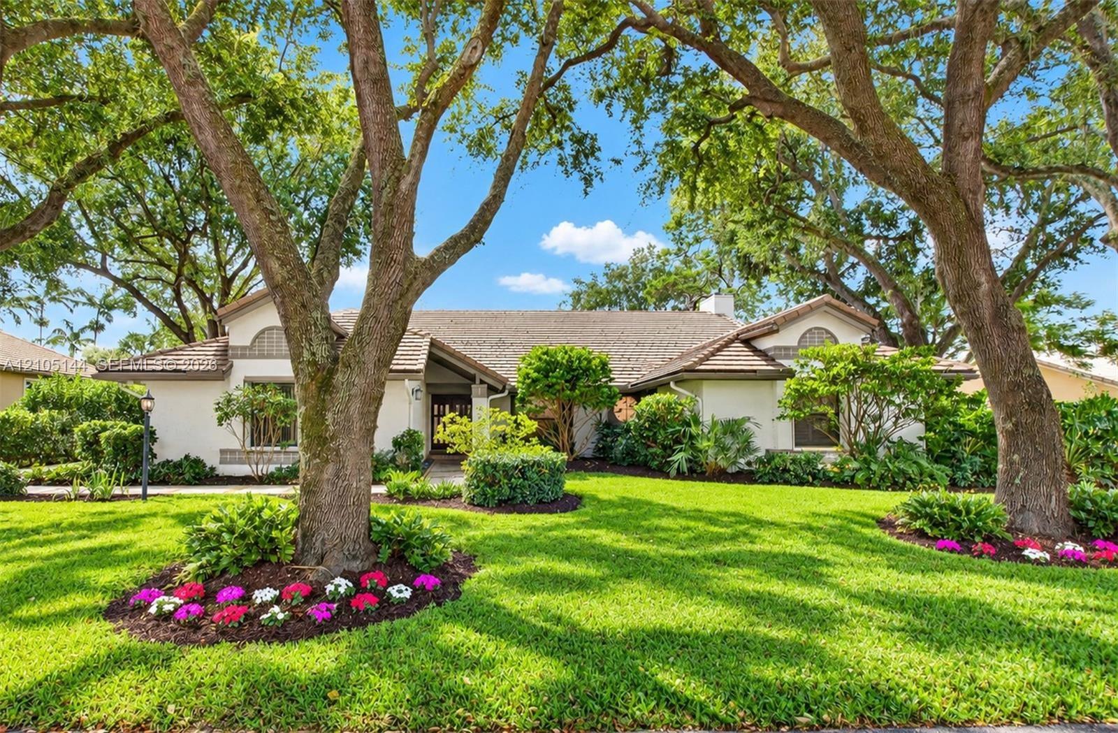 Stucco exterior featuring arched architectural details, a tiled roof, mature canopy trees, and landscaped garden beds with seasonal plantings