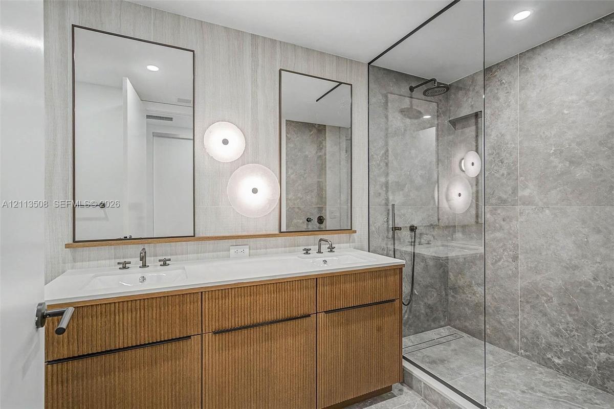 Dual-sink vanity with fluted wood-finish cabinetry, rectangular under-mount basins, wall-mounted round lighting, and a glass-enclosed shower featuring