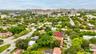 Aerial perspective of a residential property featuring a pitched roof, mature canopy trees, established landscaping, and paved street access