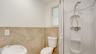 Bathroom featuring large-format beige tile wainscoting, rounded wall-mount basin, white toilet, and enclosed shower stall with built-in shelving
