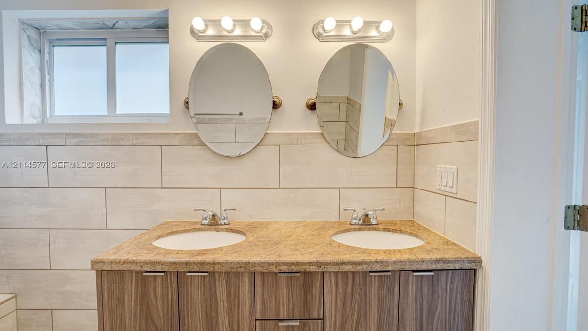Double vanity featuring a stone countertop, dual undermount sinks, wood-finish cabinetry, two oval mirrors, and horizontal tile wall surrounds