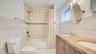 Dual-sink vanity with stone countertop, large-format tile surround, bathtub and shower combination, and oval wall-mounted mirrors