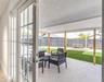 Covered patio with light-toned stone tile flooring, wooden support columns, white-framed glass doors, artificial turf, and a perimeter fence