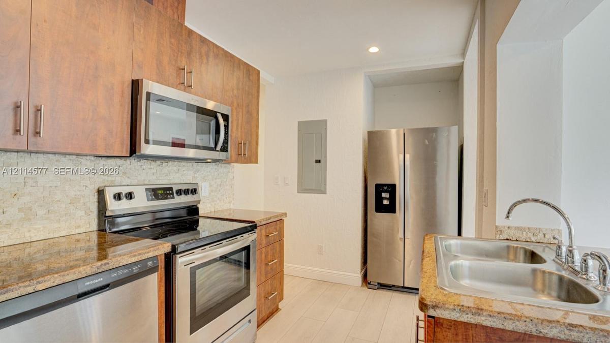 Kitchen featuring wood-finish cabinetry, stone-texture backsplash, stainless steel appliances, and solid-surface countertops with a dual-basin undermo