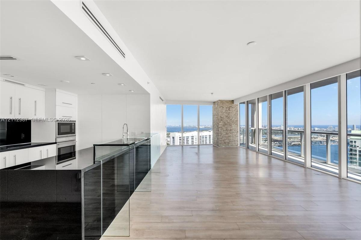 Open-concept interior featuring floor-to-ceiling windows, wood-finish flooring, a kitchen with black countertops and white cabinetry, and a stone-clad