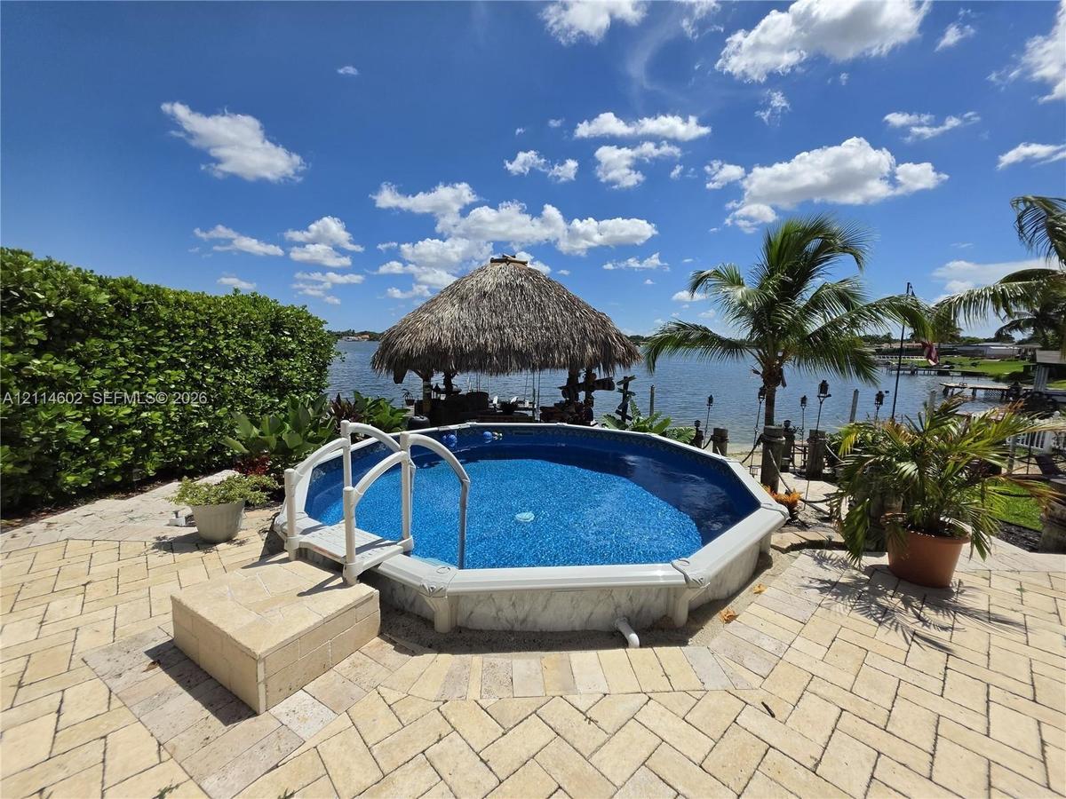Above-ground pool with a surrounding paved patio, thatched-roof cabana, and waterfront bulkhead