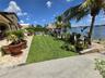 Waterfront backyard featuring a manicured lawn, stone-paved patio, mature palm trees, and a thatched-roof structure