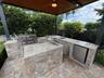 Outdoor kitchen under a covered structure featuring a natural stone-faced prep area, stone countertops, built-in stainless steel grill, undermount sin
