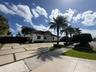 Single-story residence featuring a white stucco exterior, expansive paver driveway with turf accents, arched entryway, and mature palm trees