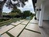 Covered patio featuring wood-finish flooring, large-format concrete pavers with turf inlays, professional landscaping, and wall-mounted exterior light