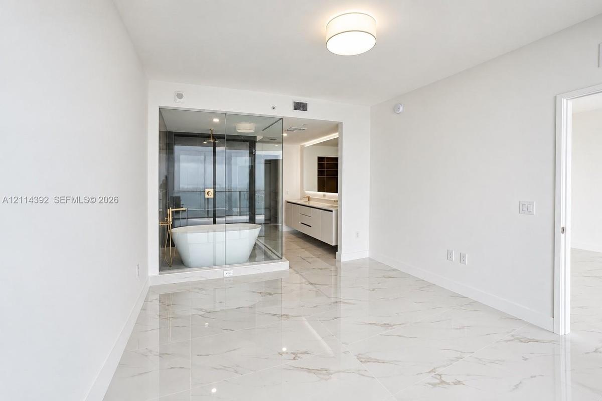 Polished stone-finish flooring, glass-enclosed bathroom area featuring a freestanding soaking tub, dual-sink floating vanity, and flush-mount lighting