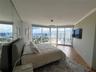Floor-to-ceiling windows, wood-finish flooring, mirrored accent wall, circular pendant lighting, and an attached balcony with water and skyline scener