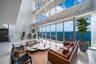 Double-height ceilings with floor-to-ceiling glass windows, full-wall stone cladding, integrated multi-pendant lighting fixture, and light-tone wood-f