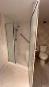 Walk-in shower enclosure featuring a handheld showerhead, integrated seating, and mounted grab bar, finished with white wall tiling and a standard toi