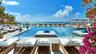 Infinity-edge pool featuring a stone-tile deck, built-in in-pool lounge platforms, glass perimeter railing, and ocean backdrop