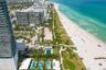 Aerial perspective of a high-rise residential building featuring multiple ground-level swimming pools, manicured palm landscaping, and direct access t