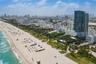 Aerial perspective featuring immediate oceanfront positioning, expansive sandy shoreline, dense coastal foliage, and high-rise urban density