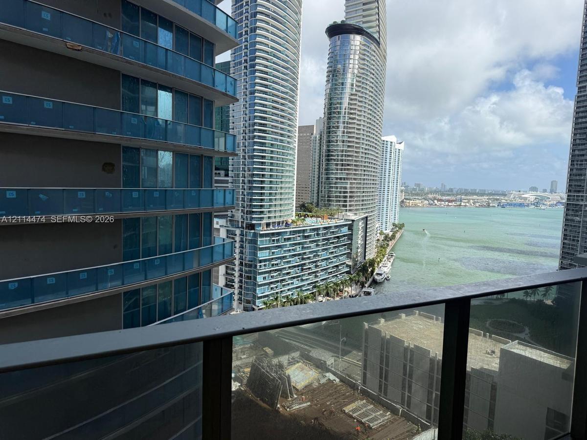 Balcony featuring metal railings with glass panels overlooking a bay, high-rise urban structures, and waterfront scenery