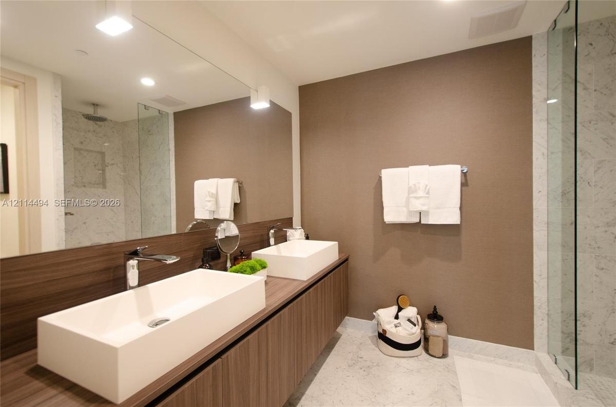 Floating vanity with dual rectangular vessel sinks, expansive wall-to-wall mirror, stone-finish flooring, and glass-enclosed shower with tiled surroun
