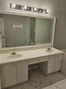 Dual-sink vanity with white cabinetry, integrated stone-finish countertop, full-width mirror, and multi-bulb vanity lighting, accented by large-format