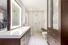 Custom dark-wood cabinetry, white composite countertops, integrated LED-backlit mirror, glass-enclosed walk-in shower with pebble-tiled wall, and buil