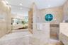 Stone-tiled bathroom featuring a jetted corner tub, walk-in shower with circular window, expansive wall-to-wall mirror, and built-in shelving