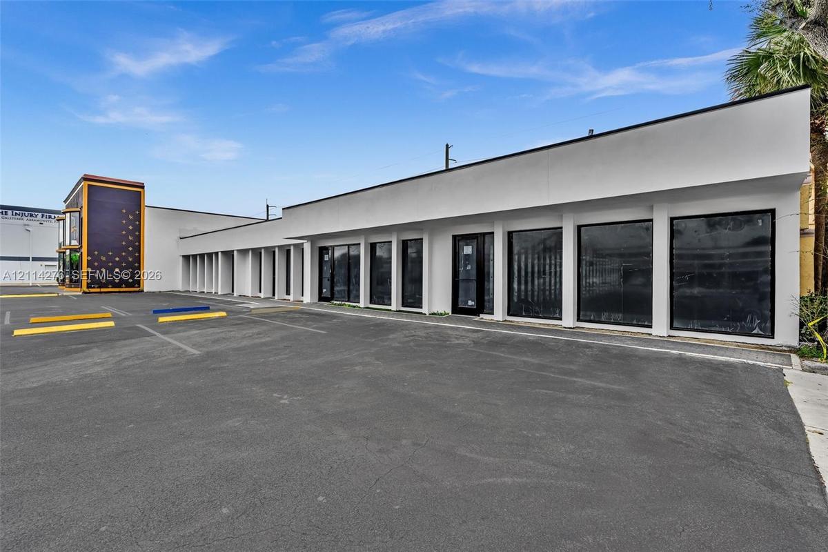 Commercial building exterior featuring a white smooth-finish facade, extensive floor-to-ceiling glass paneling, and an asphalt parking area with marke