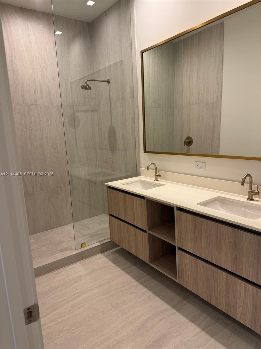 Floating dual-vanity with wood-finish cabinetry, white countertop, integrated rectangular sinks, brass-toned fixtures, and a glass-enclosed shower wit