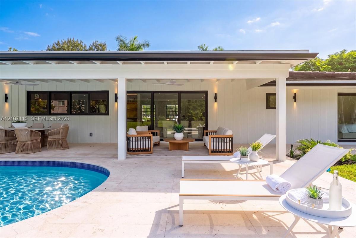 Covered outdoor patio featuring board-and-batten siding, recessed lighting, multi-panel sliding glass doors, and a stone-paver pool deck surrounding a