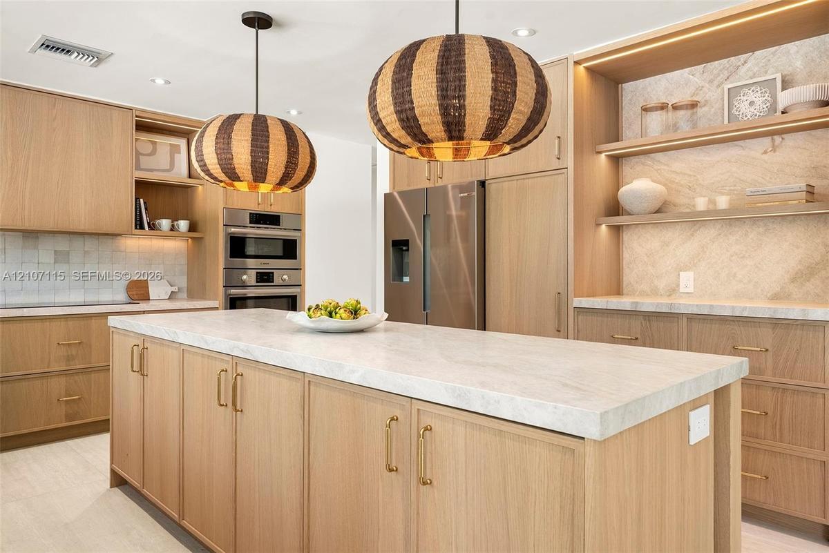 Kitchen featuring light wood-finish cabinetry, a waterfall-edge stone countertop island, built-in stainless steel appliances, decorative textured pend
