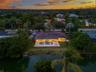 Aerial perspective of a waterfront property featuring a paved rear patio, in-ground pool, covered veranda, and mature landscaping