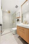 Wood-finish floating vanity with a white countertop and brass hardware, glass-enclosed walk-in shower with herringbone tile walls, and a large wall-mo