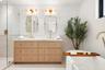 Double vanity featuring light wood cabinetry with gold-toned hardware, stone countertops, dual rectangular mirrors, and a glass-enclosed shower with h