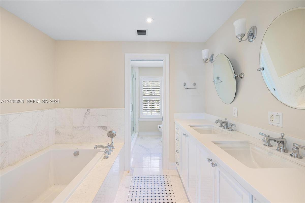 Double vanity with integrated sinks, white cabinetry, polished metal fixtures, and wall-mounted mirrors, featuring a soaking tub with stone tile surro