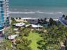 Expansive coastal grounds featuring manicured green space, mature palm trees, paved walking paths, and organic-shaped shade structures