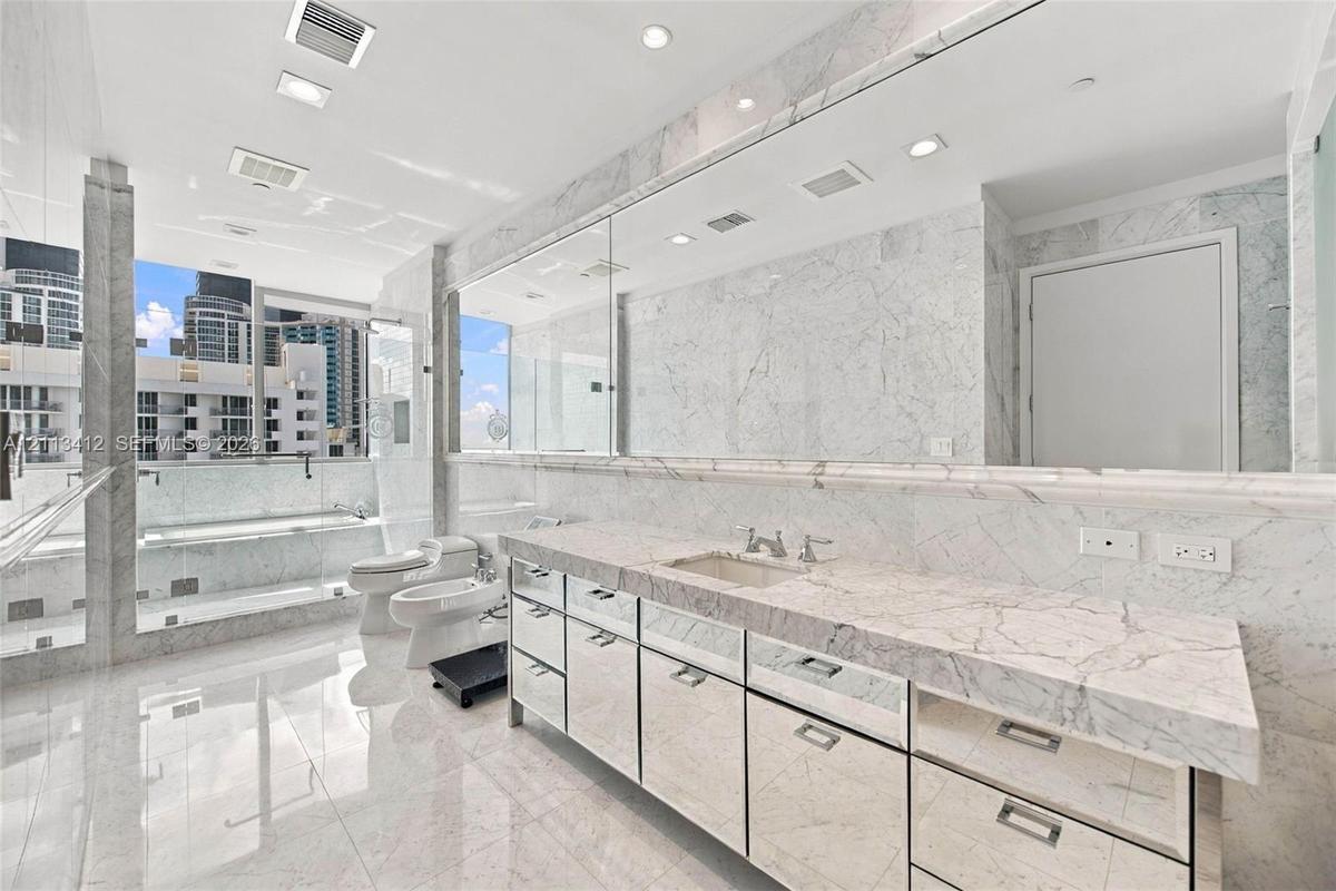 Full-length stone vanity with dual undermount sinks, expansive wall-to-wall mirror, floor-to-ceiling natural stone wall and floor tile, glass-enclosed