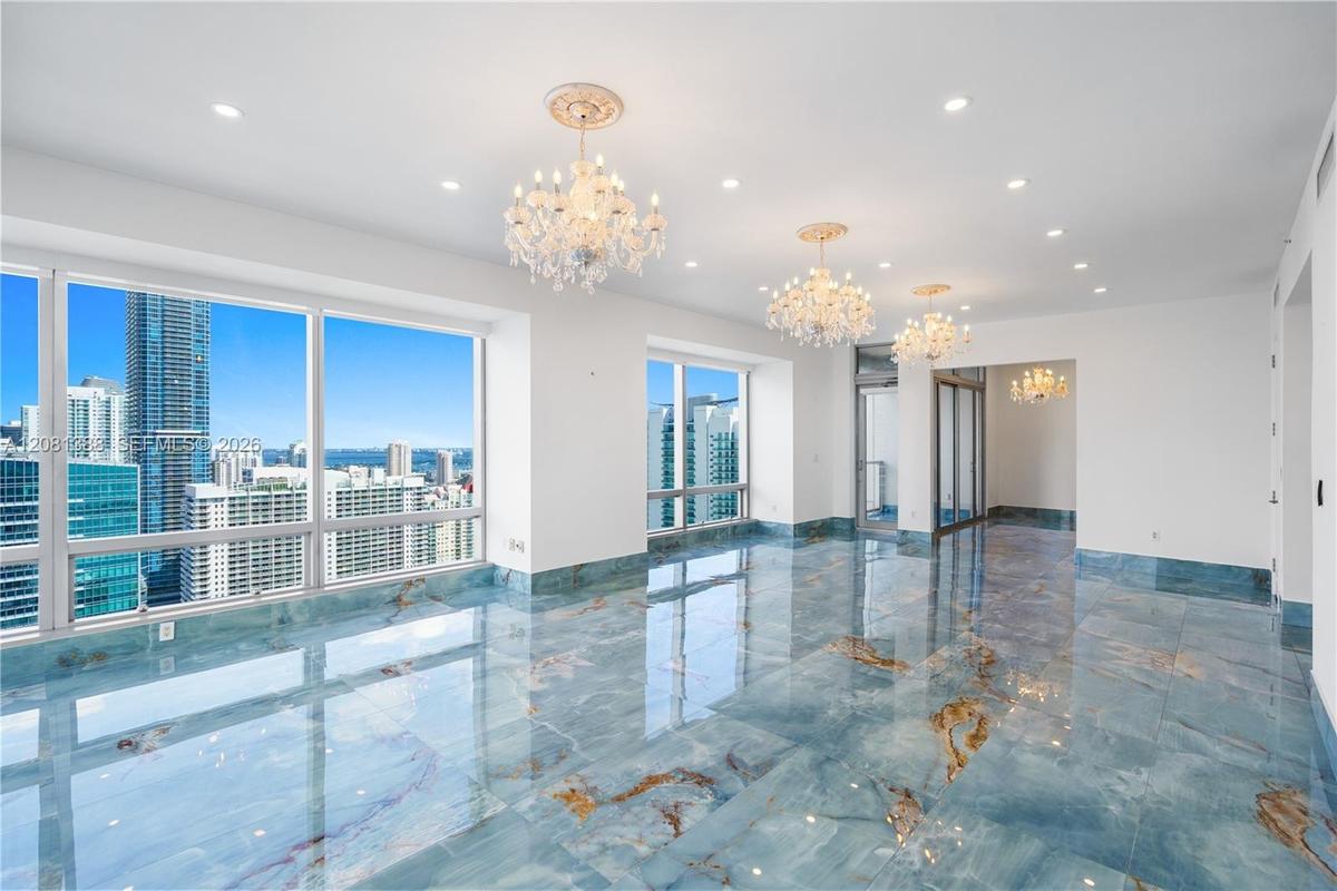 Polished stone flooring with natural veining, floor-to-ceiling windows, crystal chandeliers, recessed lighting, and glass door access to a balcony