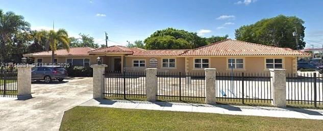 Single-story exterior featuring a clay tile roof, stucco finish, gated paved entrance, and masonry pillars