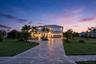 Two-story exterior featuring light-toned masonry, a multi-car garage, and an expansive paved driveway surrounded by mature palm trees and landscaped l