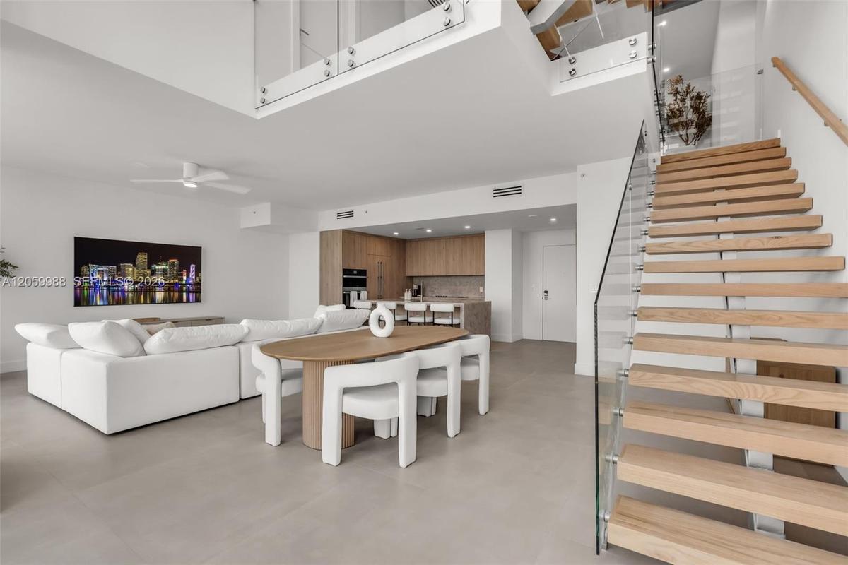 Open-concept interior featuring wood-tread staircase with glass railing, large-format neutral flooring, double-height ceiling, built-in cabinetry, and