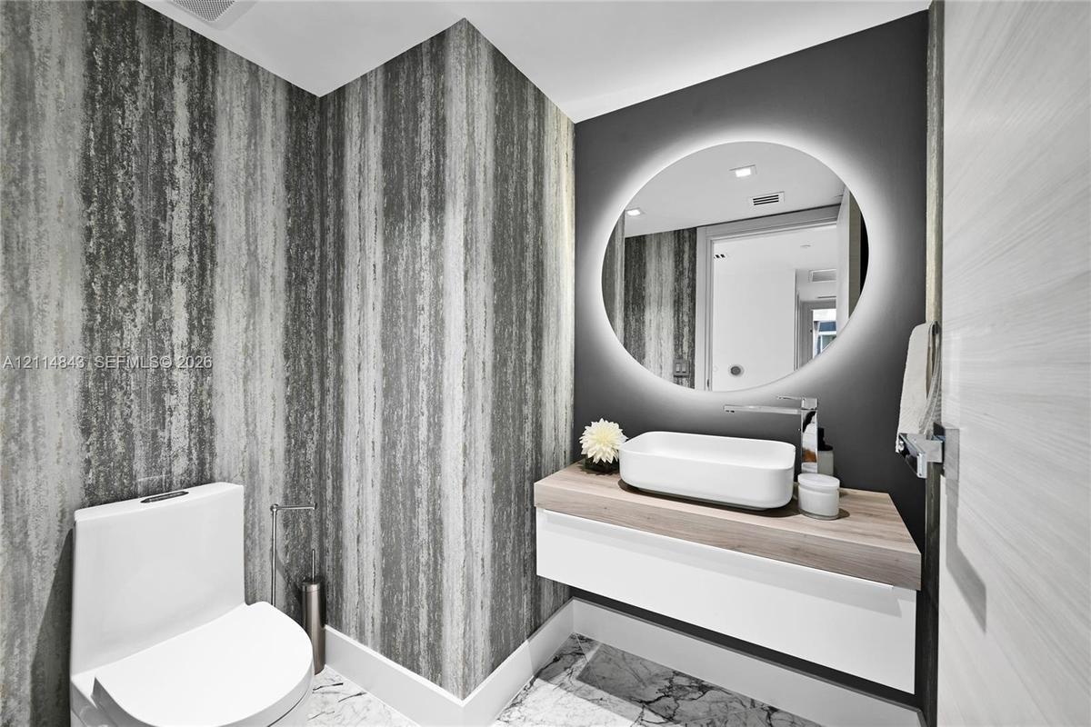 Floating vanity with wood-finish countertop, white vessel sink, polished chrome faucet, circular backlit mirror, and stone-patterned floor tile