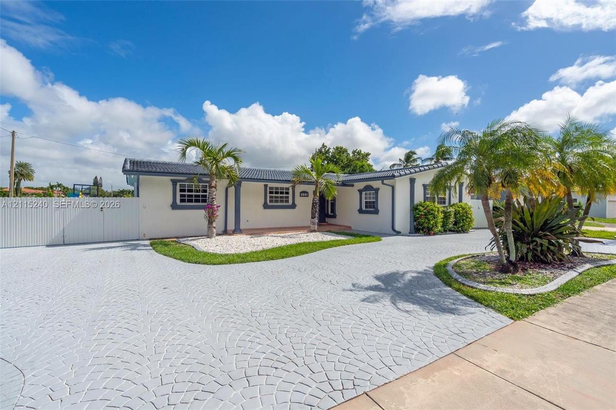 Single-story exterior featuring a decorative paved driveway, white stucco finish, dark shingle roof, integrated landscaping beds, and a white privacy 