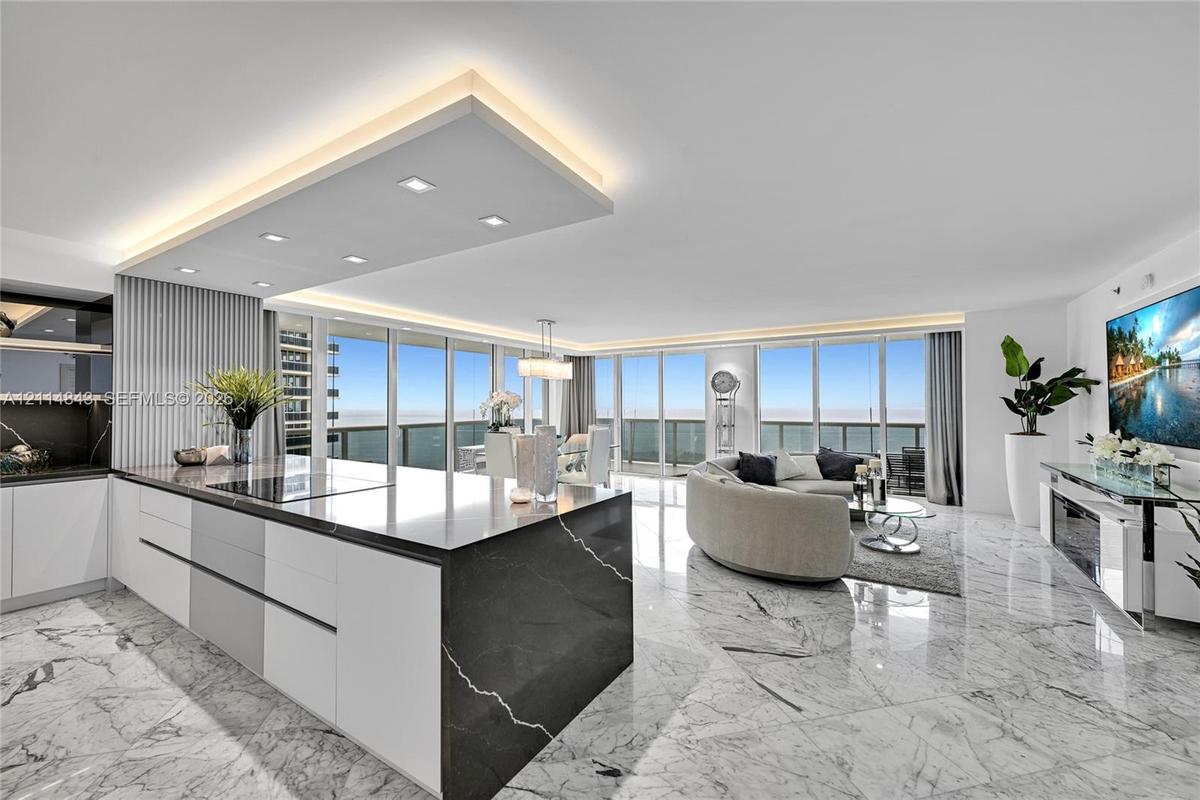 Open-concept living space featuring stone flooring, floor-to-ceiling windows, a center kitchen island with a dark stone waterfall countertop, recessed
