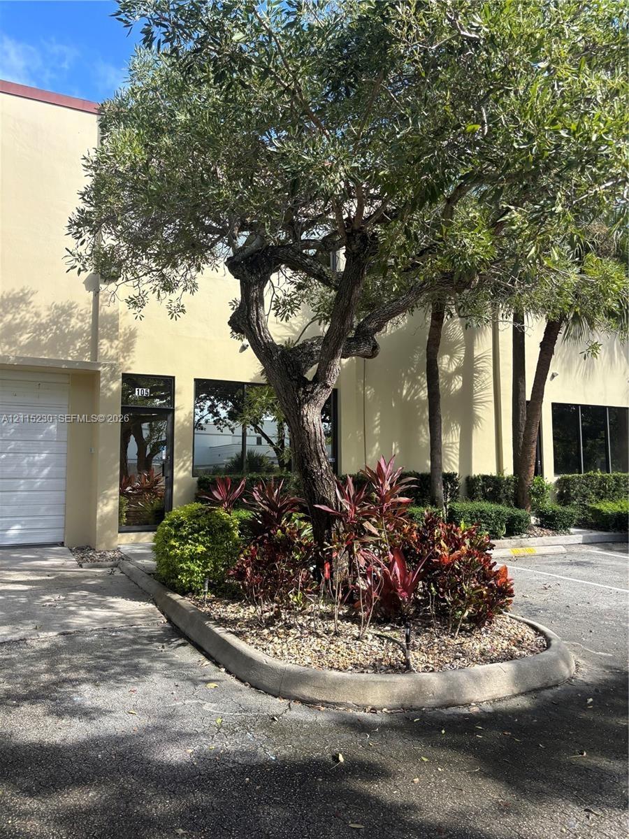 Exterior façade featuring smooth stucco finish, metal-framed windows, and a dedicated landscaped island with mature trees, shrubbery, and stone ground