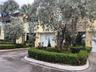 Two-story exterior featuring a light-colored stucco facade, roll-up garage doors, paved driveway, and mature landscaping including manicured shrubs an
