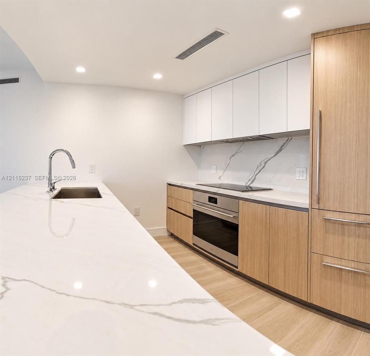 Kitchen featuring an expansive stone countertop with integrated sink, wood-finish cabinetry, white matte upper cabinets, stone backsplash with veining